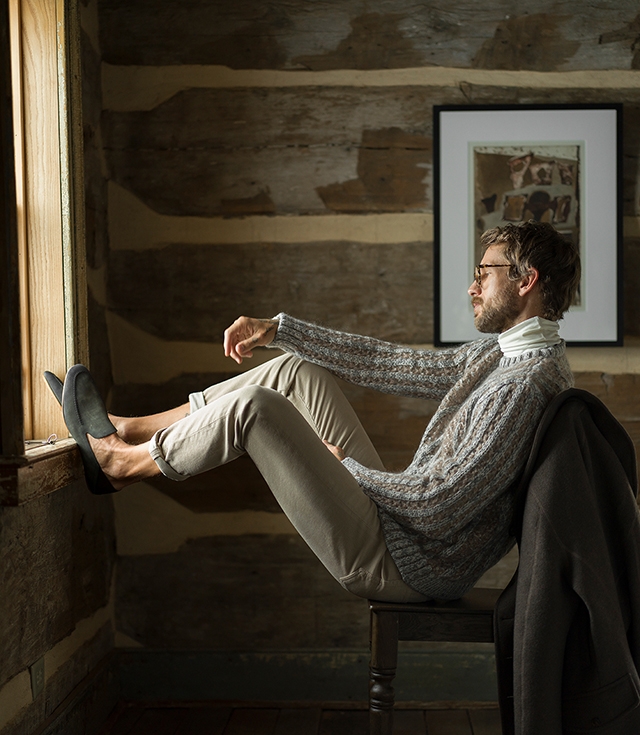 A male model sits in a cabin, wearing a sweater, tan pants, and Magnanni Heston slippers in Musgo Suede.