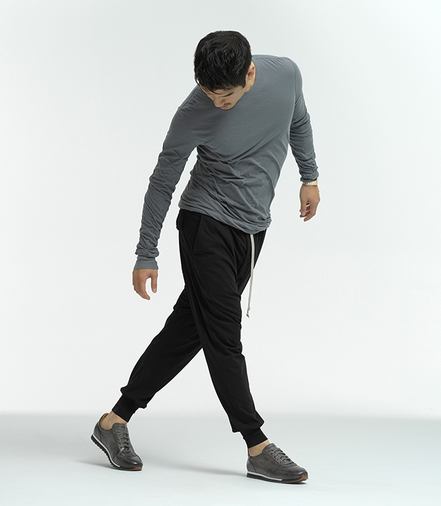 A male model awkwardly steps while wearing a grey shirt, black sweatpants, and Magnanni Ibiza sneakers in Grey.