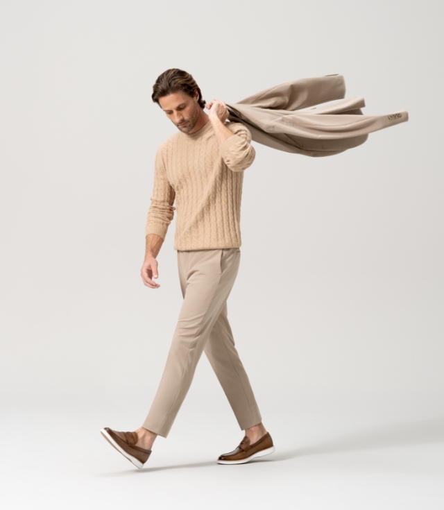 A man steps forward with such tenacity that his beige jacket is forced horizontal. He is also wearing a tan sweater, beige pants, and Magnanni Lalo loafers.
