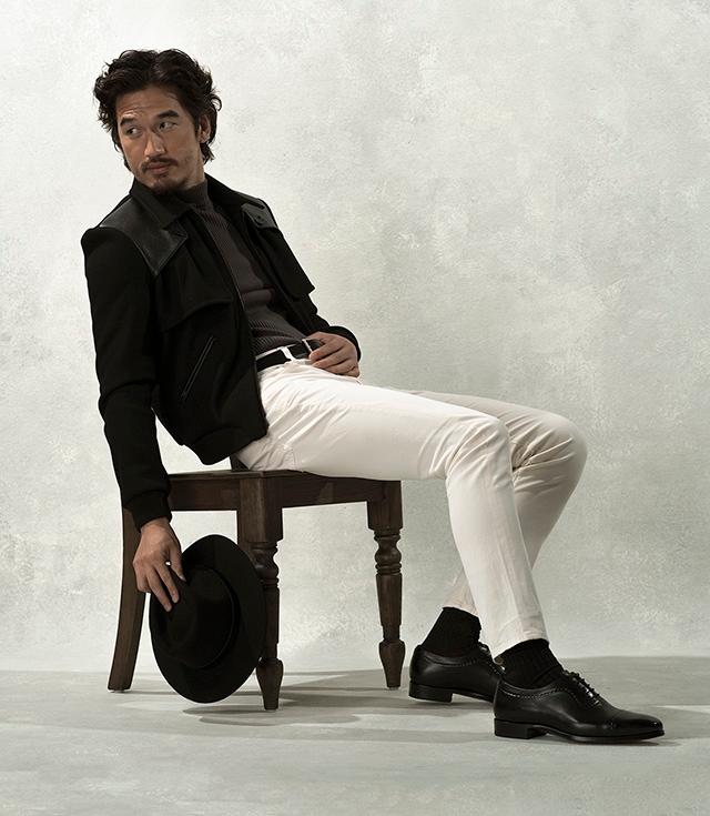 A man sits in a chair, wearing white pants, a black jacket, and Magnanni Camilo lace ups in black.