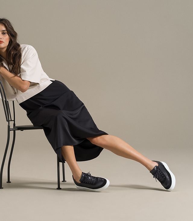 A woman leans on a chair while wearing a black skirt, white top, and Magnanni Mia sneakers in Black.