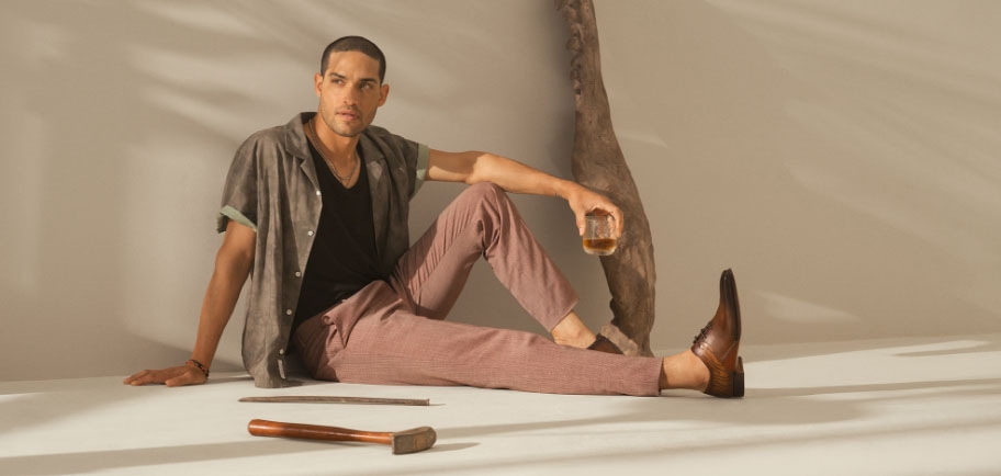 A male model sits and drinks, wearing light red pants and Magnanni Tasos Tabaco and Cuero shoes.
