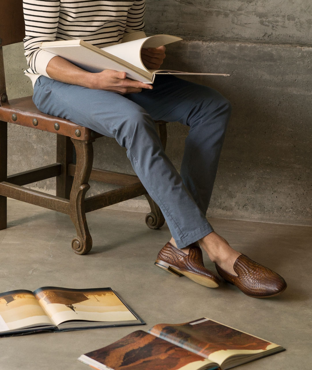 A male model wearing navy pants and Magnanni Herrera Cuero shoes reads a book while sitting on a wooden chair.